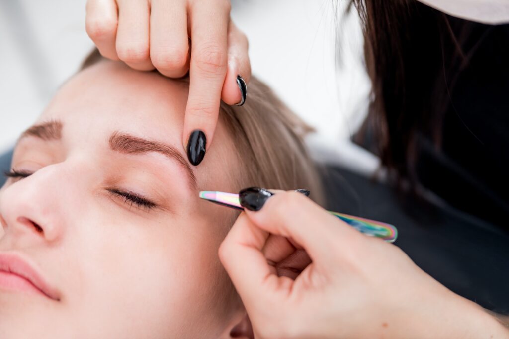 Plucking eyebrows with tweezer by beautician at beauty salon